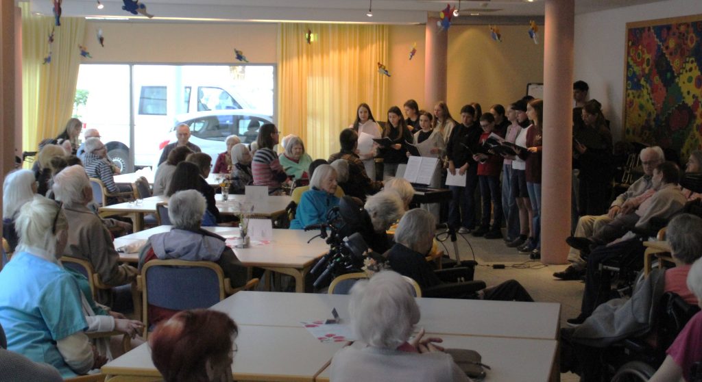 BLOG-Beitrag
FES-Schüler überraschen Bewohner im Altenzentrum musikalisch
Tränen der Rührung, Glück und Freude: So viele gute Gefühle in einem Gesicht ablesen zu können, ist wohl der beste Beweis für eine gelungene Aktion.                                                          
20 Schülerinnen und Schüler unseres FES-Jugendchors blickten bei ihrem kleinen musikalischen Ständchen im Eugen-Lederer-Altenzentrum in HDH am Mittwoch in ganz viele glückliche Gesichter: Die Schüler unseres FES-Gymnasiums und unserer FES-Realschule besuchten mit FES-Chorleiter Daniel Kindsvater und Schulleiter Andreas Borkeloh die Senioren im AWO-Altenzentrum in der Stadtmitte und überraschten mit europäischer Chormusik unterschiedlicher Stilistik aus dem aktuellen Konzertprogramm. Die Cafeteria war beim Besuch der jungen Sänger aus den Klassenstufen 7-10 proppenvoll gefüllt und die jungen Choristen erfüllten das Auditorium mit fröhlicher Musik. Besondere Freude bereitete den älteren Menschen, dass zum Abschluss des musikalischen Ständchens gemeinsam gesungen wurde. „Vielen Dank, dass Ihr heute bei uns seid. Unsere Bewohner freuen sich riesig, dass ihr für sie singt“, bedankte sich Nadine Schnell, Pflegedienstleitung im AWO-Altenzentrum am Ende des gut 30-minütigen kleinen chorischen Vortragsprogramms.
