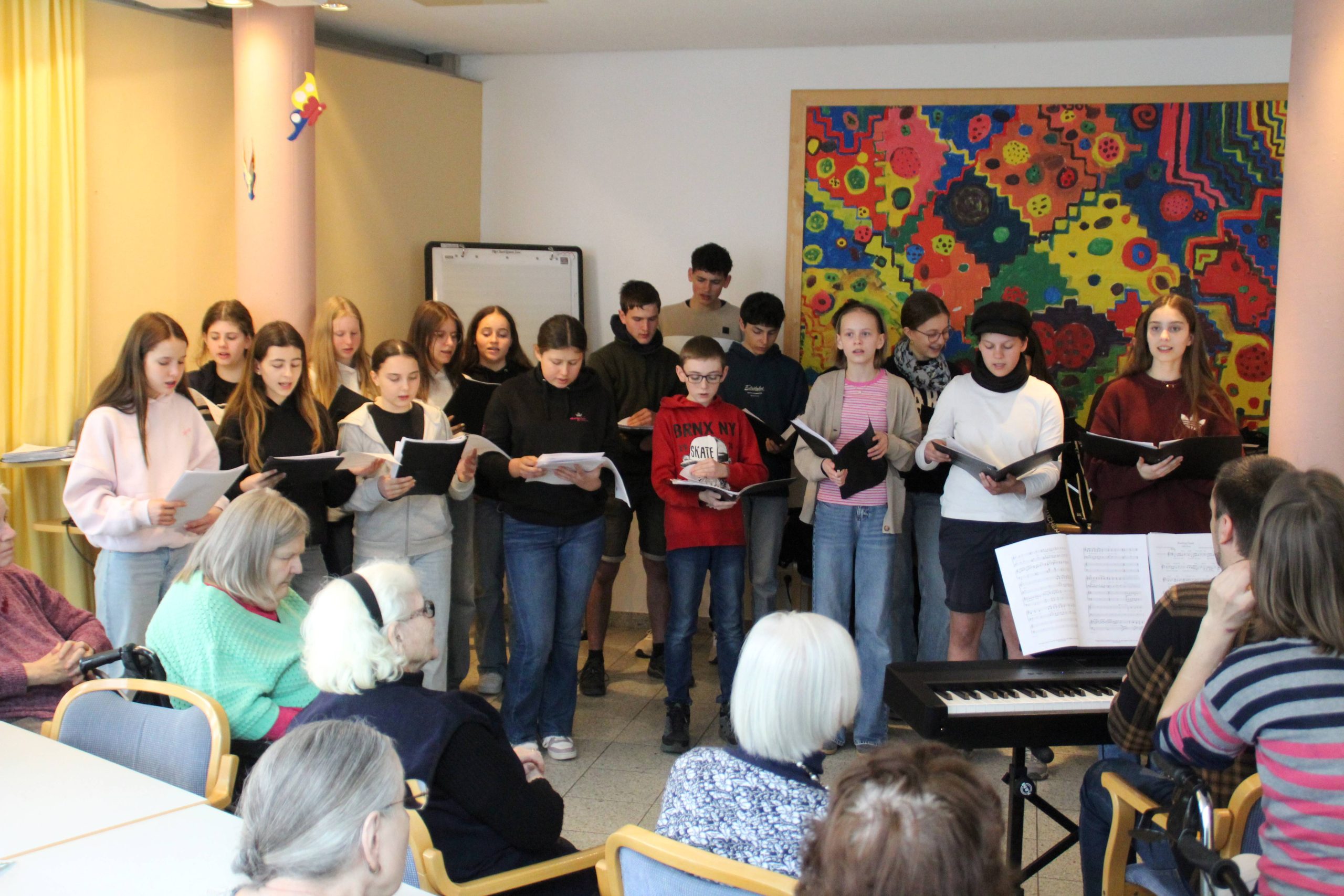 IMG_3401 BLOG-Beitrag FES-Schüler überraschen Bewohner im Altenzentrum musikalisch Tränen der Rührung, Glück und Freude: So viele gute Gefühle in einem Gesicht ablesen zu können, ist wohl der beste Beweis für eine gelungene Aktion. 20 Schülerinnen und Schüler unseres FES-Jugendchors blickten bei ihrem kleinen musikalischen Ständchen im Eugen-Lederer-Altenzentrum in HDH am Mittwoch in ganz viele glückliche Gesichter: Die Schüler unseres FES-Gymnasiums und unserer FES-Realschule besuchten mit FES-Chorleiter Daniel Kindsvater und Schulleiter Andreas Borkeloh die Senioren im AWO-Altenzentrum in der Stadtmitte und überraschten mit europäischer Chormusik unterschiedlicher Stilistik aus dem aktuellen Konzertprogramm. Die Cafeteria war beim Besuch der jungen Sänger aus den Klassenstufen 7-10 proppenvoll gefüllt und die jungen Choristen erfüllten das Auditorium mit fröhlicher Musik. Besondere Freude bereitete den älteren Menschen, dass zum Abschluss des musikalischen Ständchens gemeinsam gesungen wurde. „Vielen Dank, dass Ihr heute bei uns seid. Unsere Bewohner freuen sich riesig, dass ihr für sie singt“, bedankte sich Nadine Schnell, Pflegedienstleitung im AWO-Altenzentrum am Ende des gut 30-minütigen kleinen chorischen Vortragsprogramms.